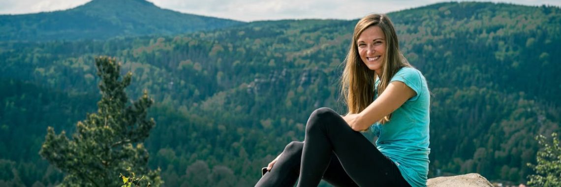 Smiling woman in blue sneakers sitting on a rock with mountain landscape in Jonsdorf, Germany.